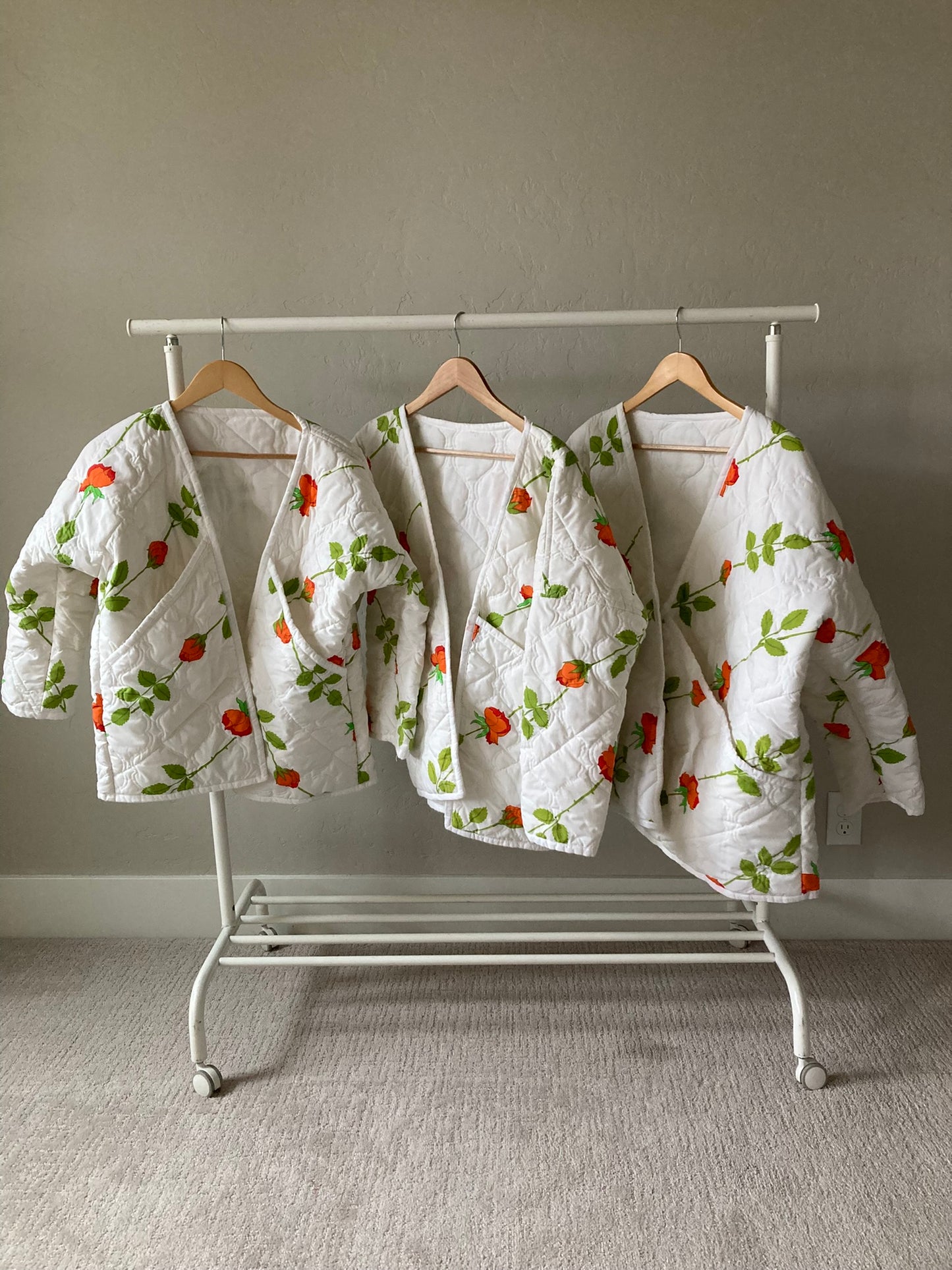 Three quilt coats hang side by side, fronts to the viewer, on wooden hangers on a white clothes rack. The coats are white with a pattern of roses going diagonally from bottom left to top right. The roses are mostly orange with some redder petals, and the stems are a bright green.