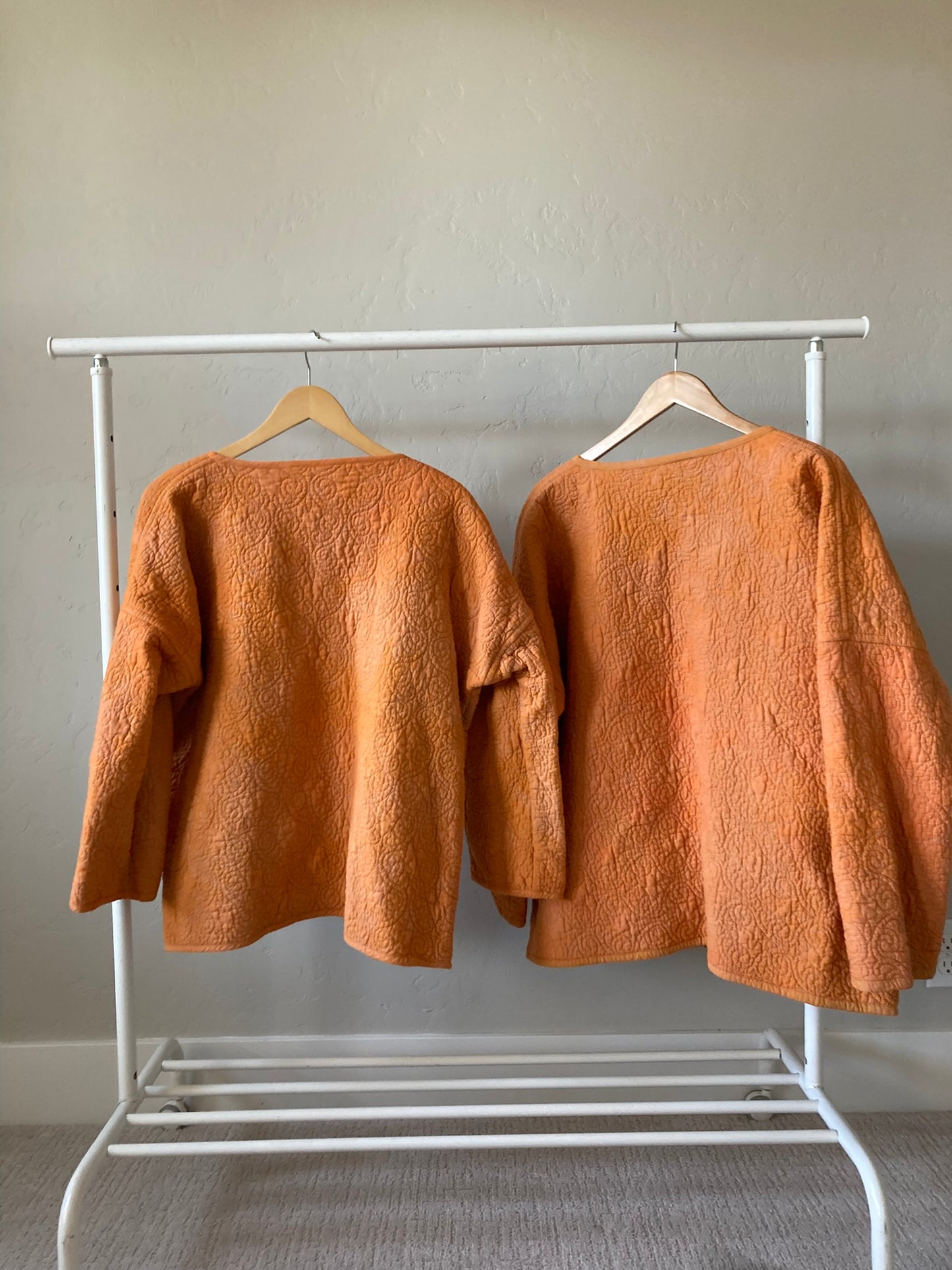 The two coats hang with the back towards the viewer, showing the scrolling quilt pattern and the slight variation in orange dye color.