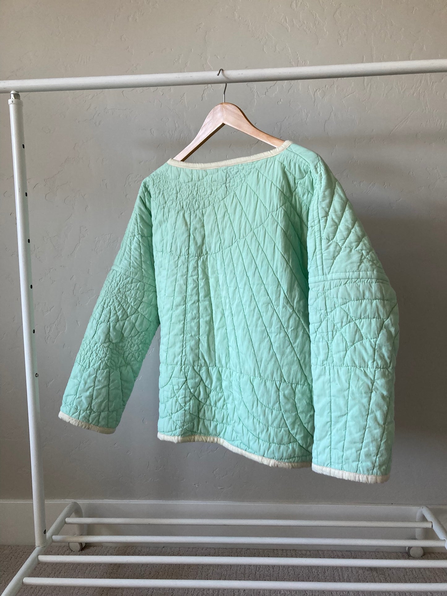 The back of the coat is toward the viewer, hanging from a wooden hanger on a white clothes rack. This view shows the and quilting froma cluster of flowers at the top left shoulder/neckline of the coat with lines radiating out from there to more concentric scalloping along the bottom of the coat. The sleeves also show, with more of the floral hand quilting and scalloping.