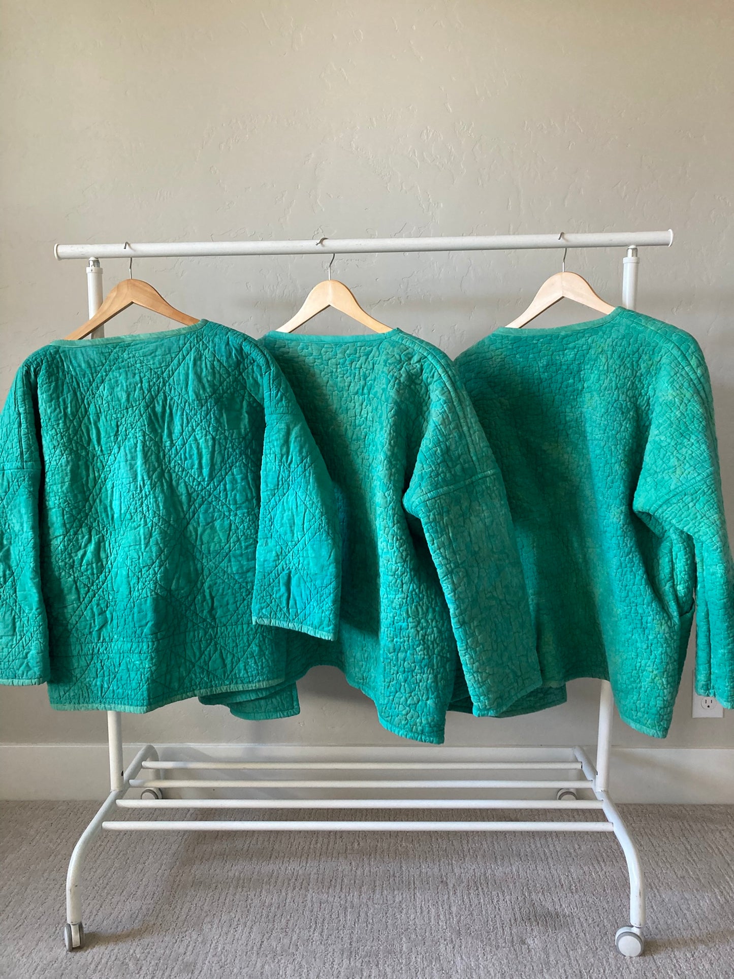 Shows the back of the three coats hanging from the clothes rack. The difference between the size small's geometric quilting and the medium/large squiggle quilting is very clear.